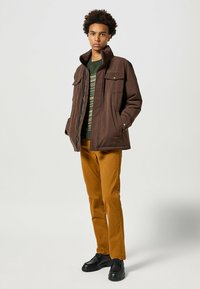 Giacca marrone con colletto alto, grandi tasche; sovrapposta a un maglione verde a fantasia. Pantaloni ramati e scarpe nere completano l'outfit.