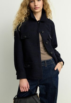 Navy blue textured jacket with a collared neckline, button front, and two chest pockets. Paired with a beige top and dark denim pants.