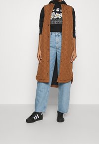 Prošívaná hnědá vesta s vysokým límcem, patentkami a bočními rozparky, vrstvená přes černou mikinu s grafickým potiskem a modré široké džíny. Černé tenisky.