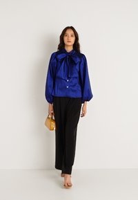 Μπλε σατέν μπλούζα με μεγάλο γιακά-κόμπο, κουμπιά μπροστά και φουσκωτά μανίκια, σε συνδυασμό με μαύρα παντελόνια. Το μοντέλο κρατά μια μικρή κίτρινη τσάντα.