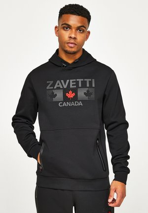 Młody mężczyzna ubrany w czarną bluzę z kapturem z napisem "ZAVETTI CANADA" oraz czerwoną ikoną klonu, stojący z jedną ręką w kieszeni.