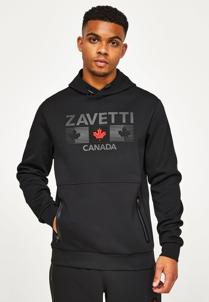 Jeune homme portant un sweat à capuche noir avec l'inscription « ZAVETTI CANADA » et un logo de feuille d'érable rouge, debout avec une main dans la poche.