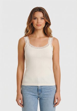 Mujer con una camiseta de tirantes blanca de tejido acanalado con borde de encaje y jeans azul claro, de pie contra un fondo liso y claro.