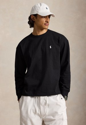 Fehér sapkát viselő férfi, fekete hosszú ujjú ing logóval a mellkasán, fehér cargo nadrág, kezek a zsebekben, oldalra néz egy egyszerű háttér előtt.