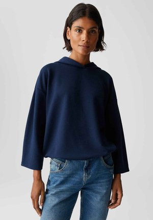 Donna con capelli scuri che indossa un maglione a maniche lunghe, blu navy, con cappuccio, e jeans di denim blu, in piedi davanti a uno sfondo grigio semplice.