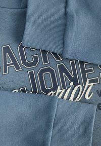 Blåt stof med en tekstureret overflade og ribbede manchetter. "Jack & Jones" trykt i store hvide bogstaver på en mørkere blå baggrund.
