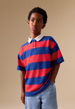 Dreng med kort krøllet hår iført en rød og blå stribet polo og lyseblå jeans, stående foran en ensfarvet beige baggrund.