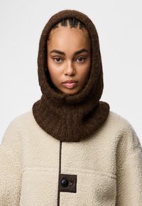 Brązowy teksturowany kaptur z dzianiny noszony na kremowej polarowej kurtce, z detalem w postaci guzika i żebrowanym wzorem.