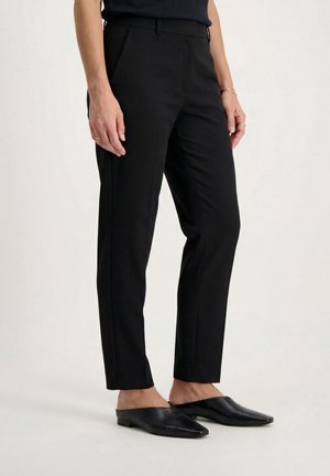 Kobieta ubrana w dopasowane czarne spodnie do kostek z bocznymi kieszeniami oraz czarne wsuwane buty z ostrym czubkiem, stojąca na białej podłodze.