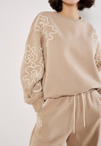 Sudadera beige con bordado floral blanco en las mangas, cuello redondo y corte relajado, combinada con pantalones joggers beige a juego que presentan acentos similares.