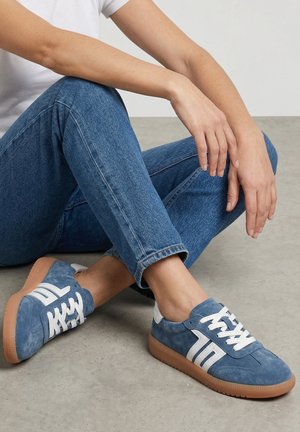 Person sidder på gulvet iført blå ruskindssneakers med hvide striber, lysebrune såler, blå jeans og en hvid kortærmet skjorte.