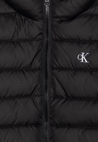 Svart boblevest med horisontale quiltede seksjoner, en front glidelås og en liten hvit "ck" logo på venstre side. Glatt tekstur.