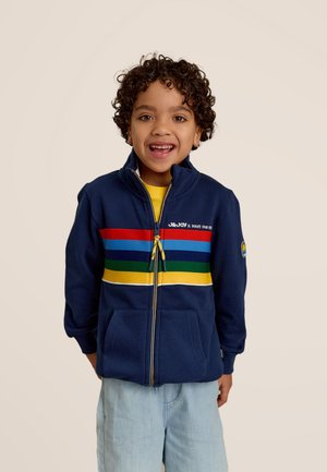 Veste zippée bleu marine avec des rayures horizontales multicolores, poches avant et col haut, associée à une chemise jaune et à un short bleu clair.