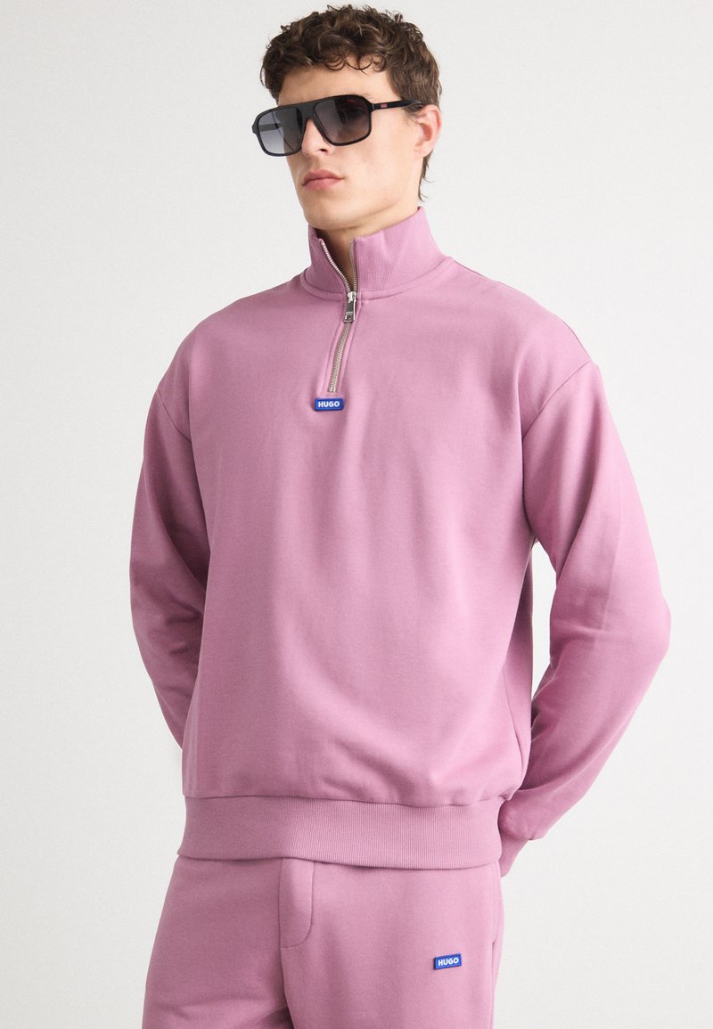 Felpa rosa a zip a un quarto in tessuto morbido, con colletto alto, tasche laterali e logo blu sul petto. Indossata con pantaloni coordinati.
