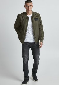 Olive green bomber jacket with a zip closure, ribbed cuffs, and pocket detail. Worn over a white t-shirt and paired with dark gray jeans and black sneakers.