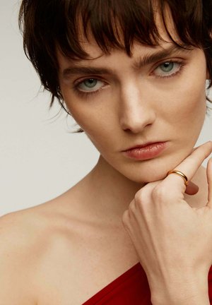 Anillo de oro en un dedo; modelo lleva una blusa roja de un solo hombro. Piel suave, fondo claro; ojos azules llamativos y maquillaje neutro acentúan las características.