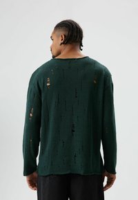 Groene trui met lange mouwen van een gestructureerde stof, met gaten van verschillende groottes en een losse pasvorm. Achteraanzicht met een ronde halslijn.