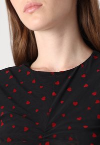 Svart tygblus med spridda röda hjärtan, rund halsringning och delikat struktur. Har en samlad detalj i mitten.