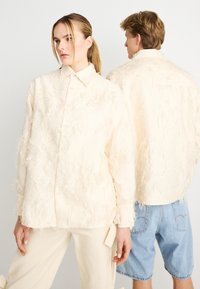 Μπεζ πουκάμισο με κουμπιά, αφράτο, με γιακά, διακοσμημένο με υφή και άνετη εφαρμογή. Φορεμένο με μπεζ παντελόνι και τζιν σορτς.