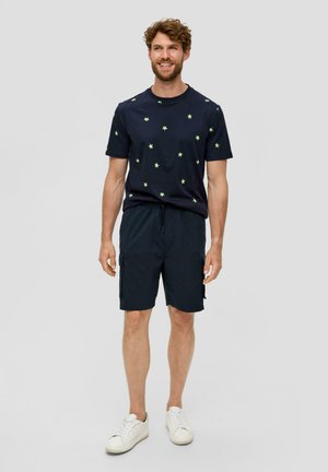 Marineblaues T-Shirt mit kleinem grünem Sternenmotiv, kombiniert mit dunkelblauen Cargo-Shorts. Modell trägt weiße Sneakers und steht vor einem neutralen Hintergrund.