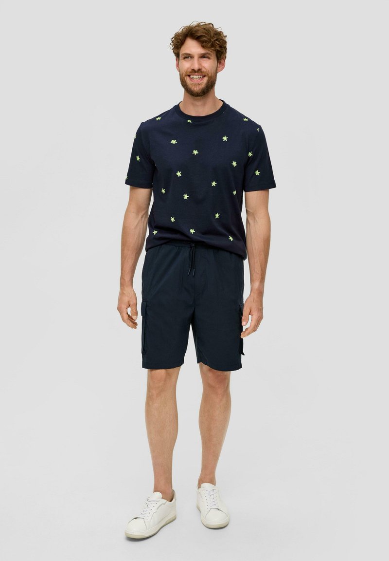 Niebieska koszulka z nadrukiem małych zielonych gwiazdek, w połączeniu z ciemnoniebieskimi szortami cargo. Model nosi białe sneakersy, stoi na neutralnym tle.