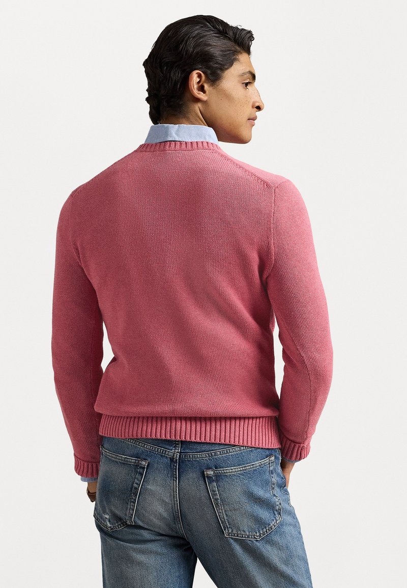 Rosa gestrickter Pullover mit rundem Halsausschnitt und geripptem Saum, getragen über einem hellblauen Hemd mit Kragen, kombiniert mit blauen Jeans.