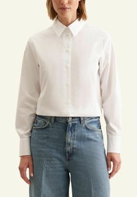 Camicia bianca con colletto classico, maniche lunghe e texture liscia, abbinata a jeans in denim blu chiaro.