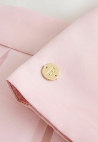 Tela de color rosa claro con una textura suave, que incluye un botón circular dorado con un detalle en forma de "B". La tela está doblada y superpuesta de manera ordenada.