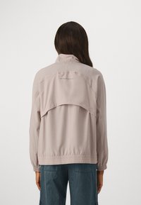 Veste de sport rose clair avec un col haut, une taille élastique et des manches froncées. Présente un logo contrastant dans le haut du dos.