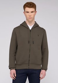 Olivgrön zip-hoodie med en texturerad yta, dragsko på huvan och en liten logotyp på bröstet. Kombinerad med mörkblå jeans.