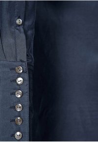 Camicia blu navy con bottoni, caratterizzata da una texture liscia, sette bottoni metallici lungo la chiusura e dettagli delle maniche leggermente ripiegati.