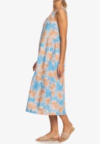 Robe maxi à fleurs bleues arborant des motifs de feuilles de palmier orange, design sans manches, tissu avec une finition légèrement texturée, accompagnée de sandales beiges.