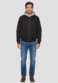 Giacca bomber nera con cappuccio grigio, chiusura a zip, tasche laterali, polsini e orlo a costine. Indossata con jeans blu e scarpe marroni.