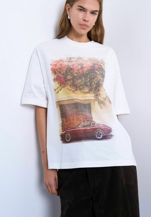 Unge kvinde iført en hvid, oversized T-shirt med et tryk af en vintage rød bil og blomstrende vinranker på fronten.