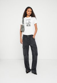 Λευκό γραφικό t-shirt με μαύρο κείμενο, γκρι παντελόνι cargo με τσέπες, μαύρες μυτερές γόβες και υφασμένη γκρι τσάντα.