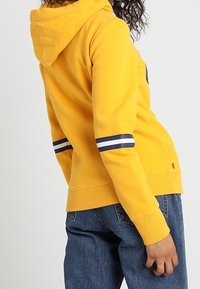Persona vistiendo una sudadera amarilla con rayas marino y blancas en la manga, combinada con unos jeans de mezclilla azules, de perfil.