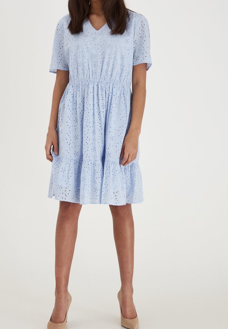 Femme portant une robe bleu clair à manches courtes en V avec un motif en œillets et un ourlet à volants, associée à des talons hauts beiges, debout.