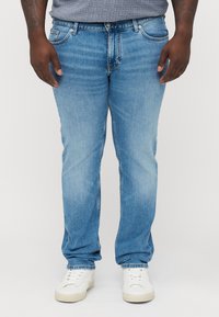 Man som bär ljusblå jeans, vita sneakers och en grå skjorta, stående med avslappnade armar vid sidorna mot en enhetlig bakgrund.