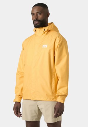 Homme portant une veste de pluie à capuche jaune Helly Hansen et un short beige, debout devant un fond gris clair uni.