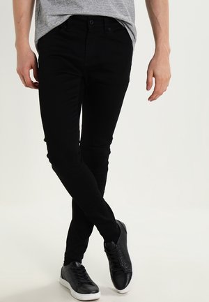 Personne portant un pantalon noir ajusté et des baskets noires à semelles blanches, debout jambes croisées devant un fond uni.
