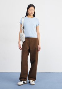 Camiseta azul clara com logotipo preto, calças largas castanhas e ténis brancos com detalhes pretos. Mala cruzada cinza com fecho.