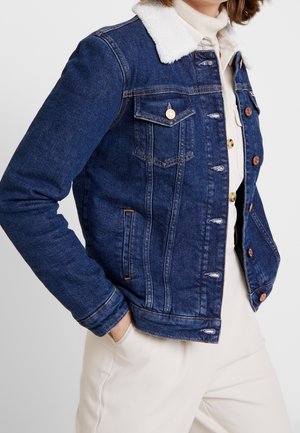 Niebieska kurtka dżinsowa z białym kołnierzem z sherpa, noszona na kremowej koszuli i spodniach, ręce w kieszeniach kurtki.