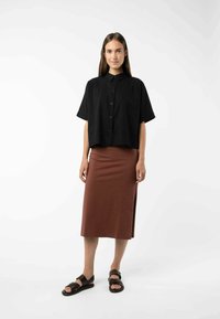 Zwarte cropped knoopshirt, bruine rok tot de knie met een zijsplit, en bruine sandalen. Soepele, lichte stof; eenvoudig ontwerp.