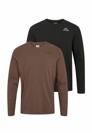 Homme debout, les bras croisés, portant une chemise marron à manches longues, un pantalon noir à jambes larges et des baskets blanches sur fond gris clair.