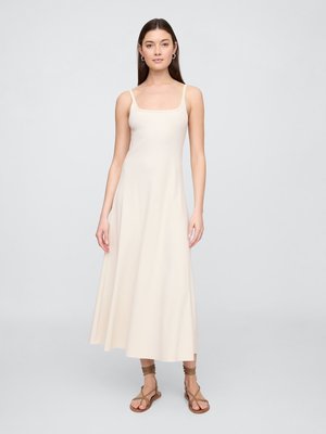 Vrouw met steil bruin haar, gekleed in een mouwloze, crèmekleurige midi-jurk en bruin leren sandalen met bandjes, staand tegen een effen grijze achtergrond.