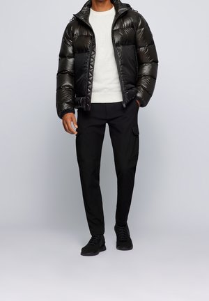 Mies, jolla on musta kiiltävä toppatakki, valkoinen neulepaita, mustat cargo-housut ja mustat lenkkarit, seisoo yksinkertaisen harmaan taustan edessä.