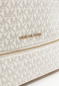 Michael Kors tas in textuur crèmekleurige stof met een herhalend logopatroon. Voorzien van een gouden logo plaat en gouden ritsdetails.