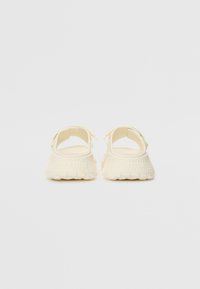 Lyse kremfargede sandaler med åpen bak, strukturert såle og myke stropper. Designet har en formet konstruksjon og minimal detaljer.