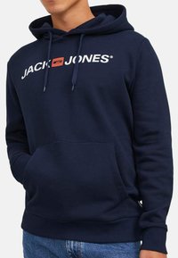 Tummansininen puuvillahuppari, jossa on etutasku, säädettävä naruhuppu ja valkoinen "JACK & JONES" -logo rinnassa.