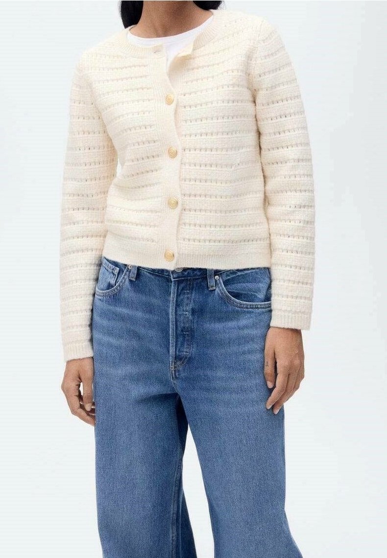Femme portant un cardigan tricoté de couleur crème avec des boutons dorés sur un haut blanc et un jean en denim bleu taille haute.
